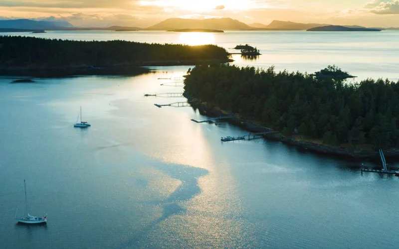 Aerial view of a coastal landscape during sunset with calm water, sailboats, a forested shoreline, and distant mountains.