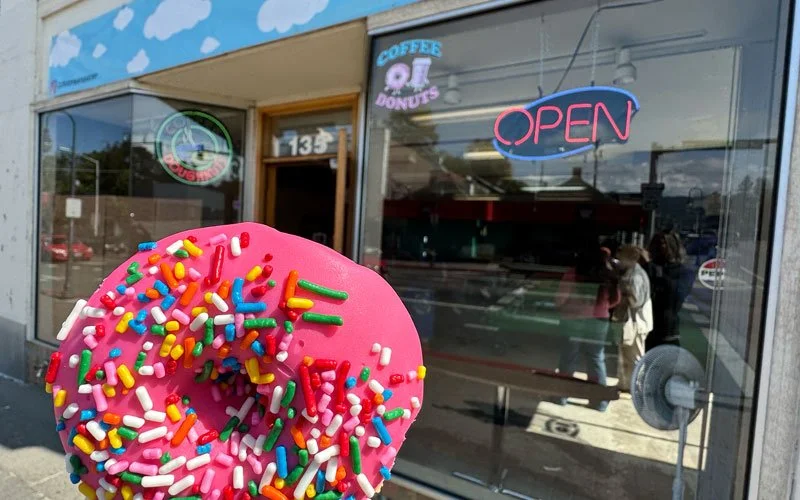 Close-up of a pink frosted donut with colorful sprinkles held in front of a donut shop window with neon 'OPEN' sign and signs for coffee and donuts