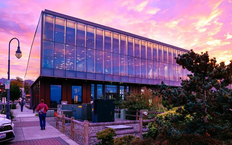 Modern glass building reflecting colorful sunset sky, surrounded by trees and people walking on the sidewalk.