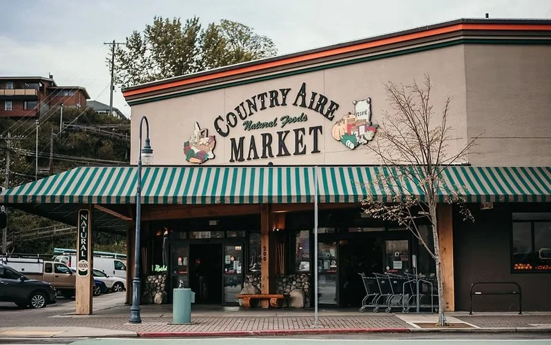 Exterior of a market named 'Country Aire Market' with a green and white striped awning, parking lot with cars, leafless tree, and a building on a cloudy day.