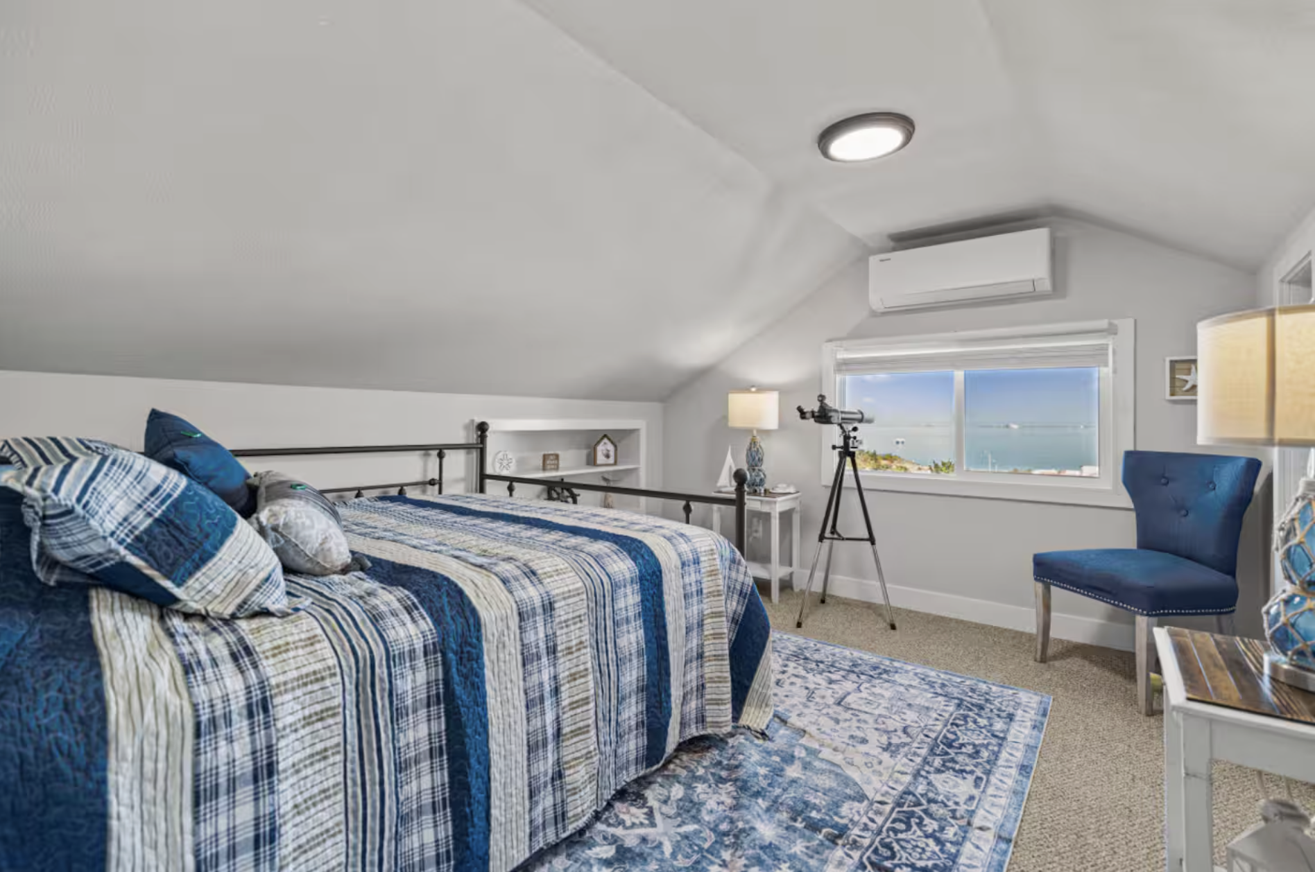 A bright attic bedroom with a blue and white color scheme, featuring a bed with a plaid quilt, a large window overlooking the ocean, a telescope near the window, and a blue upholstered chair.