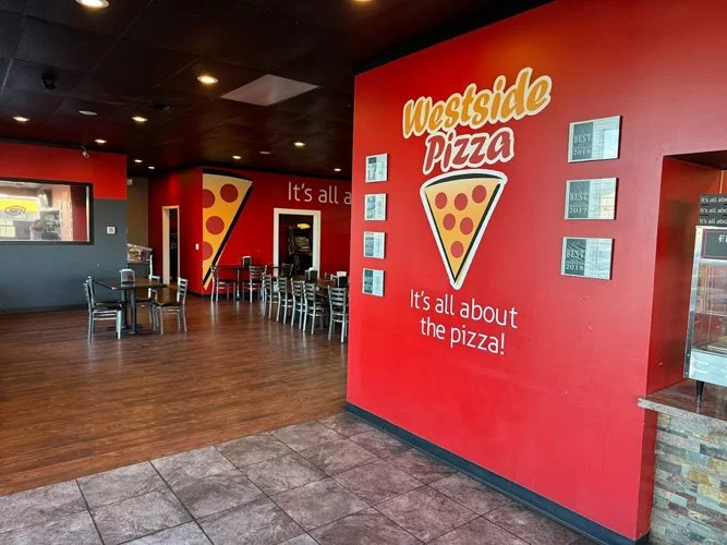Interior of a pizzeria with red walls, a large pizza graphic, and the text 'Westside Pizza, It's all about the pizza!'. There are chairs and tables inside, and a window on the left.