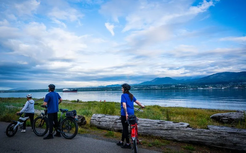Three people with bikes and helmets enjoying a scenic view by a lake with mountains in the background and a cloudy sky.