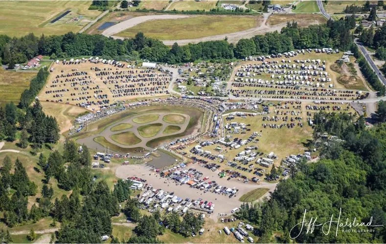An aerial view of a large outdoor parking lot filled with cars, surrounding a small pond with islands and wildlife area, and bordered by trees and grassy fields.