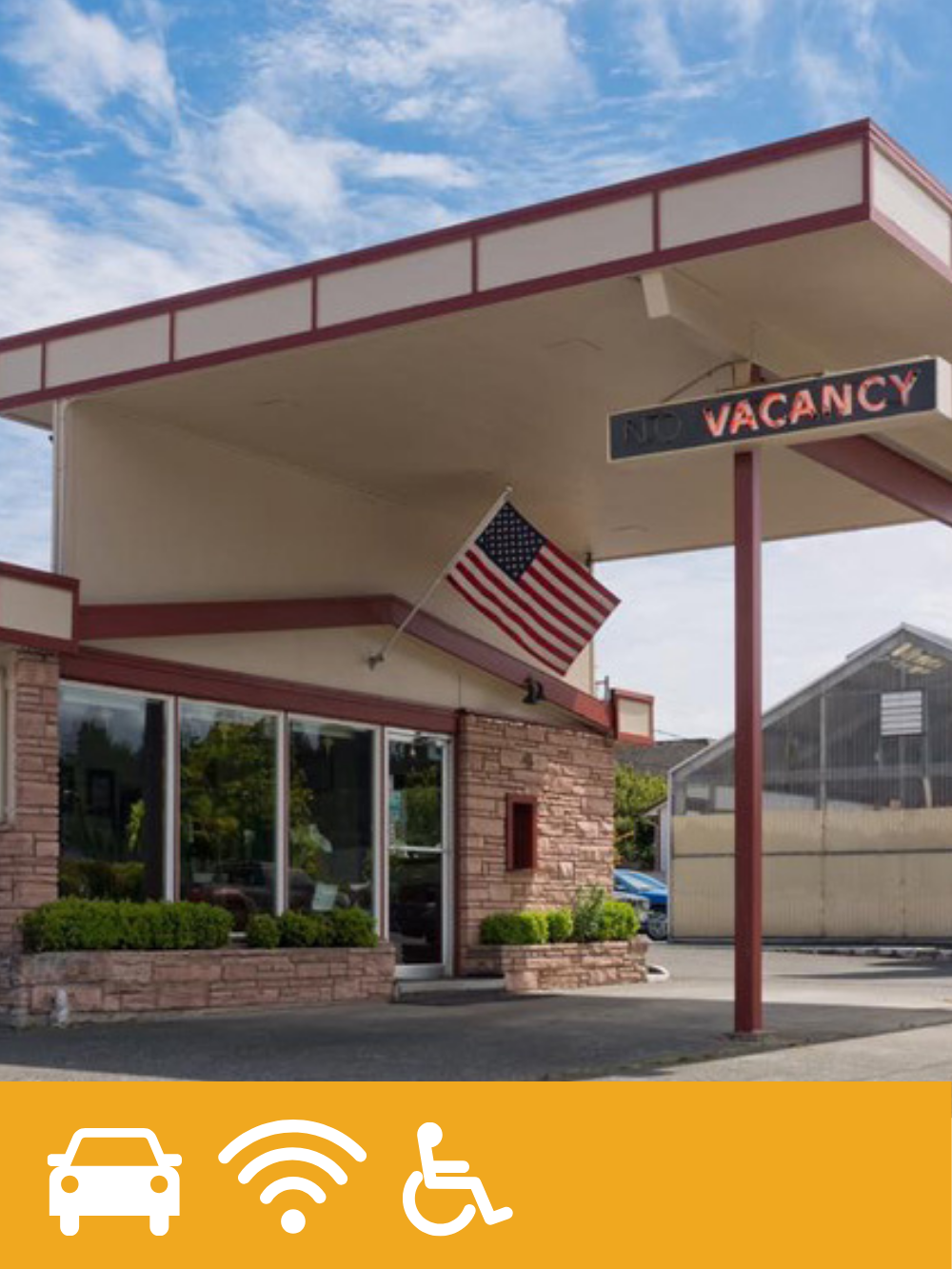 A small building with a brick facade, large windows, and a green hedge in front, with a canopy extending over the sidewalk. An American flag is mounted on the building, and a sign reads 'NO VACANCY'.