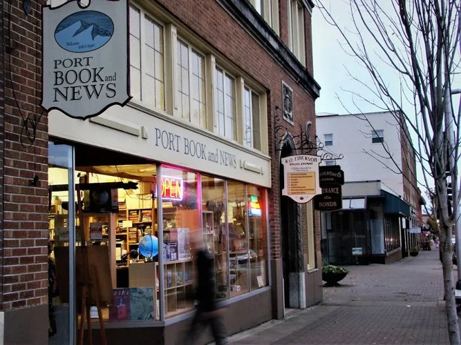 Street view of a bookstore called Port Book and News with a glass storefront, neon 'OPEN' sign, and several other signs hanging on the building, in a small town neighborhood.