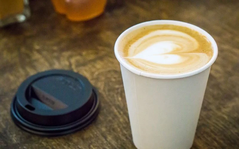 A white paper cup of coffee with latte art on a wooden table, next to a black plastic lid for the cup.