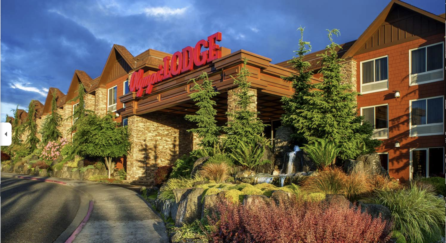 Exterior view of the Royal Lodge hotel with lush greenery and a small waterfall in front, under a partly cloudy sky in the evening.