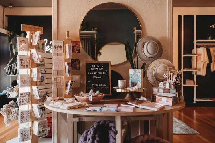 A decorative round mirror on a wooden dresser with hats, plants, jewelry, and greeting cards displayed around it, in a boutique store setting.