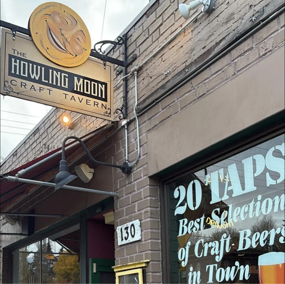 Sign for Howling Moon Craft Tavern with a smiling crescent moon logo, brick building exterior, and window displaying a menu offering 20 taps of craft beer in town.