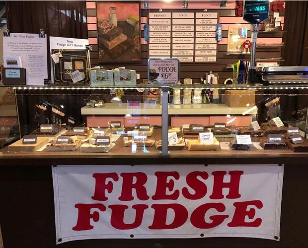 A display case filled with various types of fudge for sale, with a bright red and white banner reading "FRESH FUDGE" hanging below. Behind the case, there are signs and labels, a scale, and shelves with additional items.