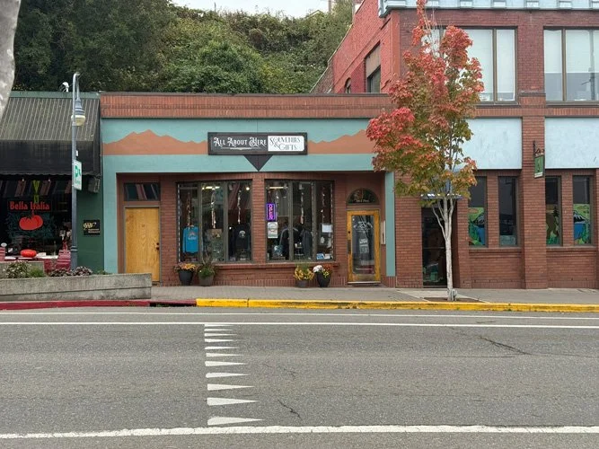 Street-facing storefront with a mountain landscape painting above it, signage reading 'All About Dogs' and 'Schnauzers & Cats,' large front windows, two flower pots, and a small tree beside the building.