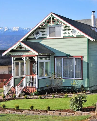 A light blue, Victorian-style house with decorative trim, a covered porch, and a front yard with lush green grass and small shrubs, with mountains in the background.