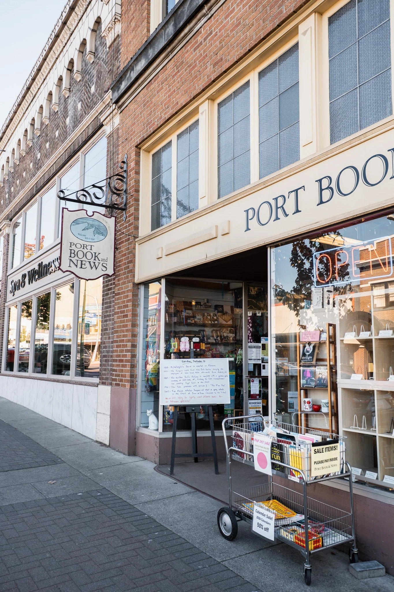Street view of a bookstore called Port Book and News with a glass storefront, neon 'OPEN' sign, and several other signs hanging on the building, in a small town neighborhood.