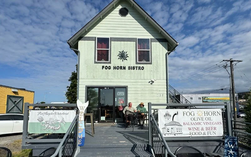 Two people sitting outside the Fog Horn Bistro on a wooden deck with signs advertising wine, beer, balsamic vinegars, and olive oils, in front of a two-story green building with red curtains and a sun logo on the wall.
