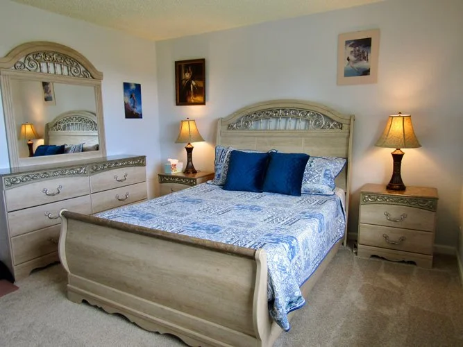 A bedroom with a wooden bed frame, blue patterned bedspread, and matching pillows. Two bedside tables with lamps, a dresser with a large mirror, and framed pictures on the walls.