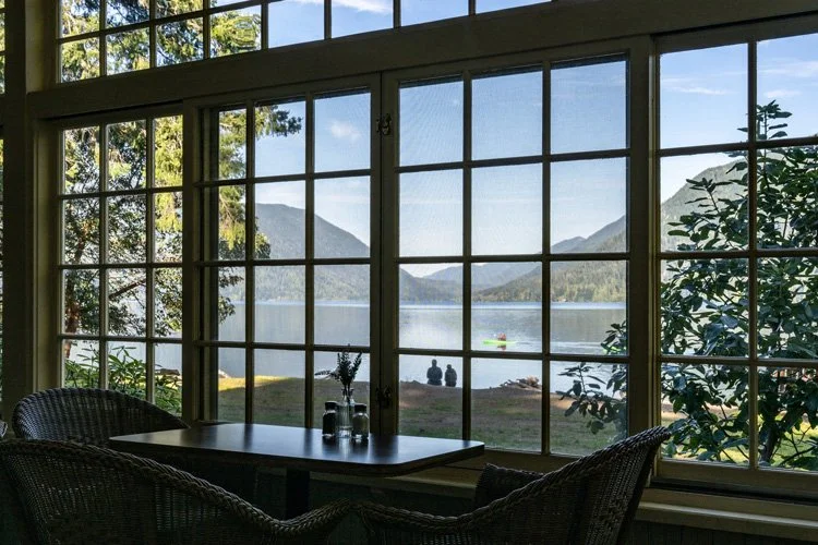 A view through a large window shows a lake with mountains and two people sitting by the water, with a kayak floating nearby. Inside, there is a table with bottles and two wicker chairs.