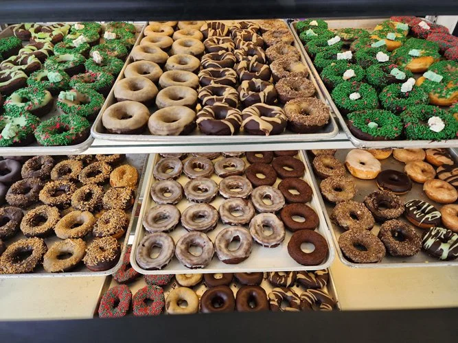 Display case filled with various types of decorated donuts arranged on trays, with colorful sprinkles, icing, and toppings.