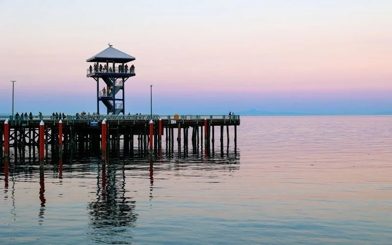 A pier with a small gazebo and railing extends into calm water, with a distant horizon and colorful sunset sky.