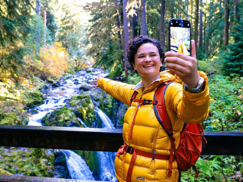 A woman in a yellow jacket taking a selfie with a smartphone in a forest with a waterfall in the background.