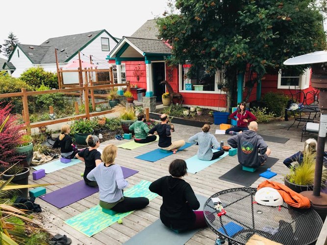 People participating in a yoga class outdoors on a wooden patio surrounded by plants and a colorful house with a tree in the background.