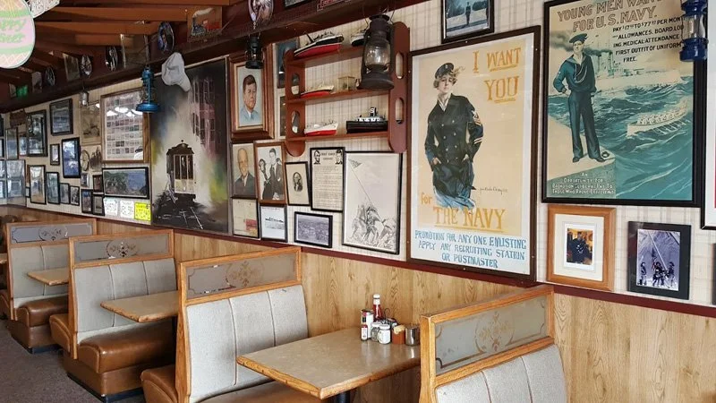 Interior of a vintage-style restaurant or cafe with wooden booths and walls decorated with navy-themed posters, framed pictures, and memorabilia.