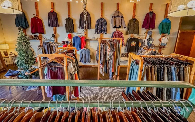 Interior of a clothing store displaying various shirts, sweaters, and pants on racks and wall hooks, with a Christmas tree and lamps in the background.