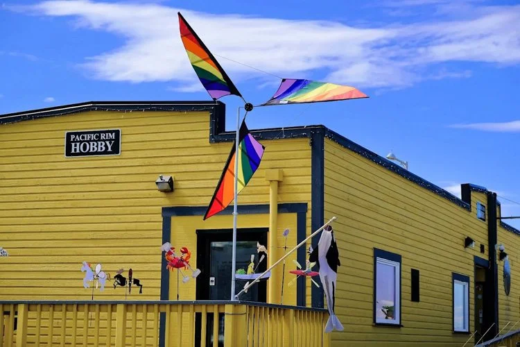 Colorful rainbow pinwheel spinning in front of a yellow building with signs reading 'Pacific Rim Hobby', decorated with various hanging decorations.