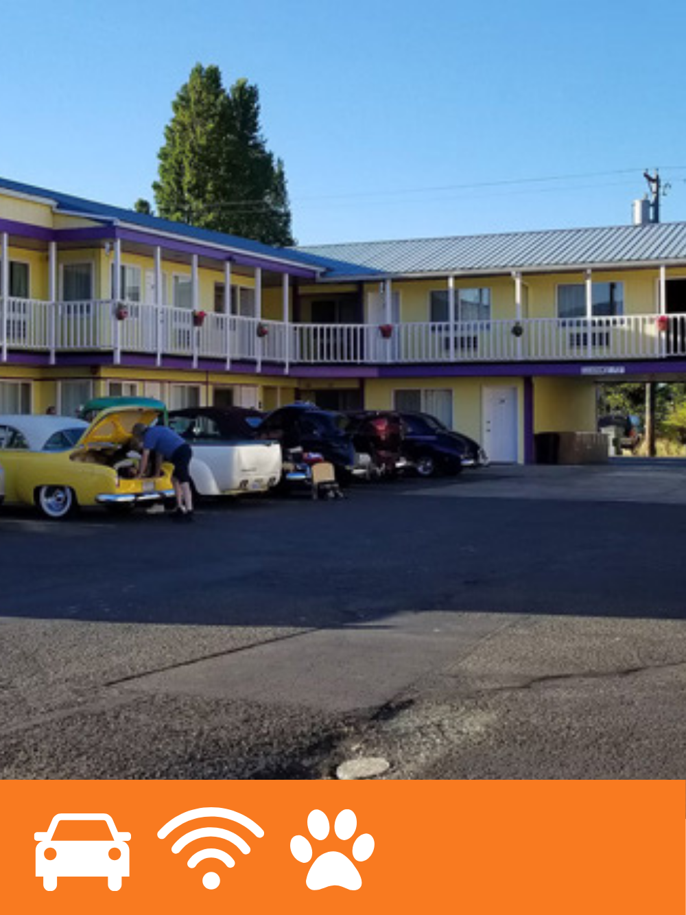 A man working on a yellow vintage car with its hood open in front of a two-story yellow apartment building with purple and white accents, several cars in the parking lot, and a tall tree against a clear blue sky.