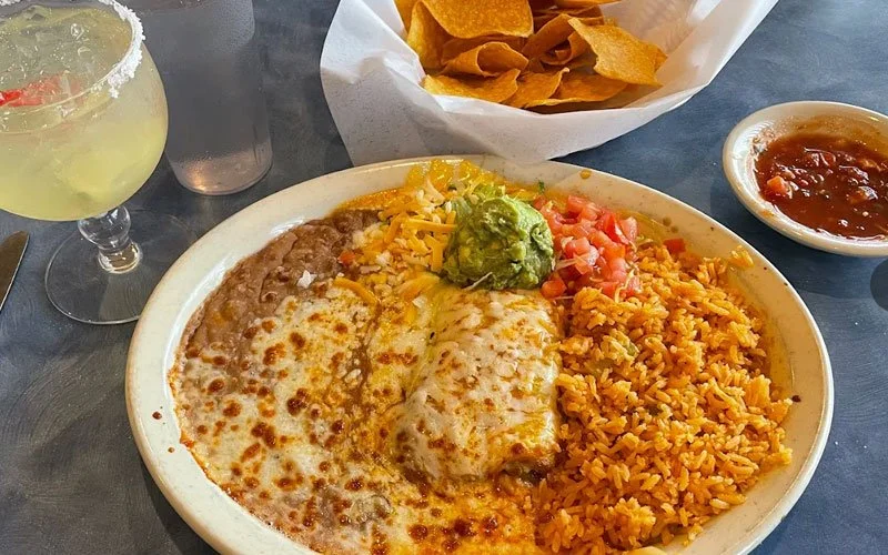 Plate of Mexican food with cheese-covered enchilada, Mexican rice, guacamole, and diced tomatoes; side of salsa and nachos; drink with lime; on dark table.