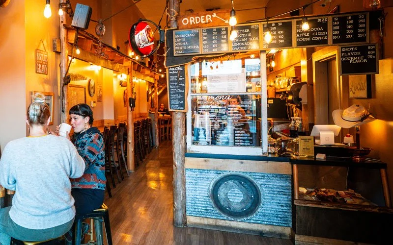 Interior of a cozy cafe with warm lighting, two women sitting at a table near the wall, a chalkboard menu, and a counter with an order window.