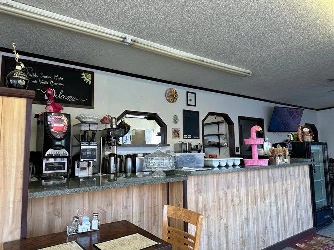 Coffee and snack bar in a small cafe with a black menu board, coffee machines, a pink flamingo water dispenser, cups, and a television mounted on the wall.