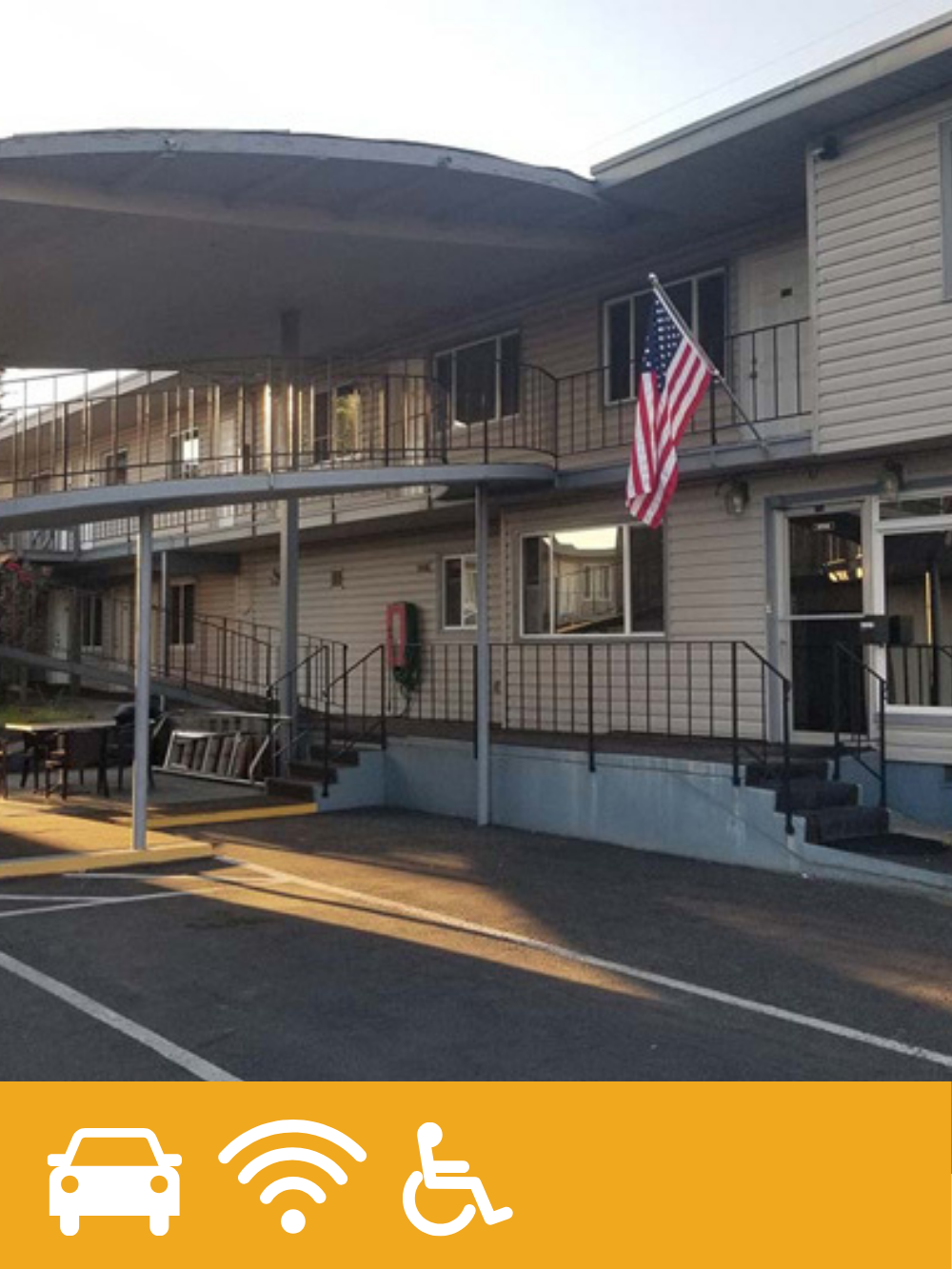 Two-story apartment building with beige siding, black railings, and a U.S. flag on a pole outside. There is an American flag hanging on the balcony, a fire extinguisher near the entrance, and a parking lot in front.
