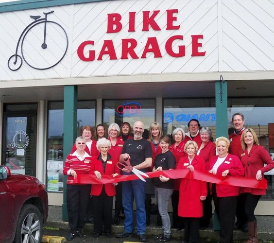 Group of people at a ribbon cutting ceremony in front of a bike repair shop called 'Bike Garage.' They are standing outside near a red car, some wearing red jackets, and holding large scissors cutting the ribbon. The shop has an 'OPEN' sign and a bicycle graphic in the window.