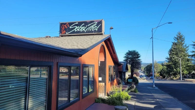 A restaurant with a sign reading 'Sabor Thai' on top, a brown exterior, large windows with blinds, and a sidewalk in front. Trees and street lamps are visible along the road.