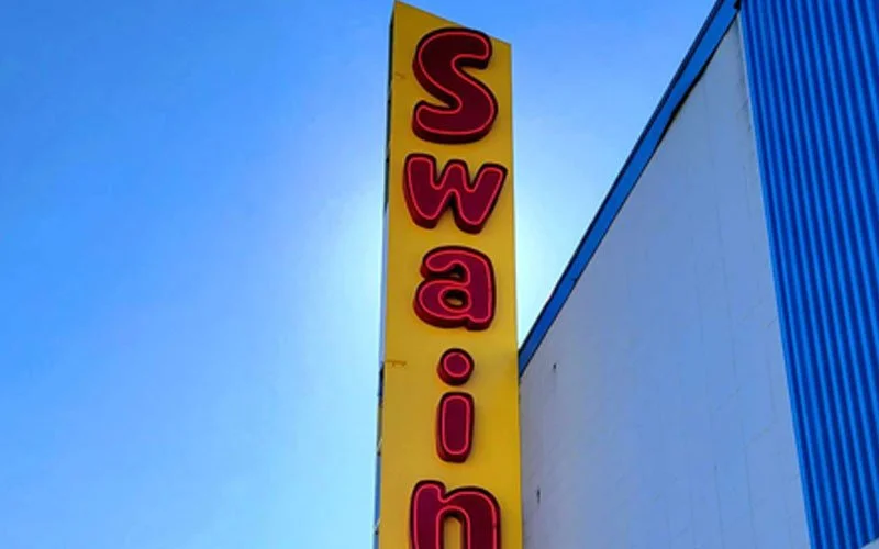 Vertical yellow sign with red letters spelling "Swain" against a blue sky and part of a blue building.
