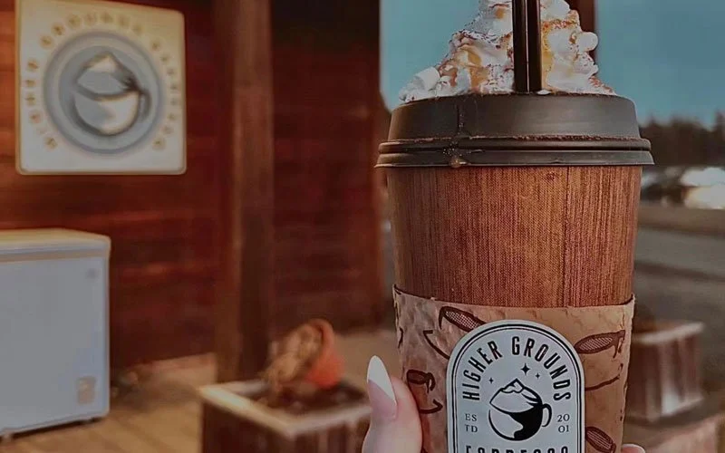 A large iced coffee or espresso drink in a to-go cup with whipped cream and caramel drizzle, held outdoors near a wooden building.