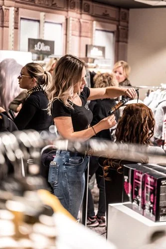 A hairstylist flat-ironing a client's hair in a busy salon with other clients and staff in the background.
