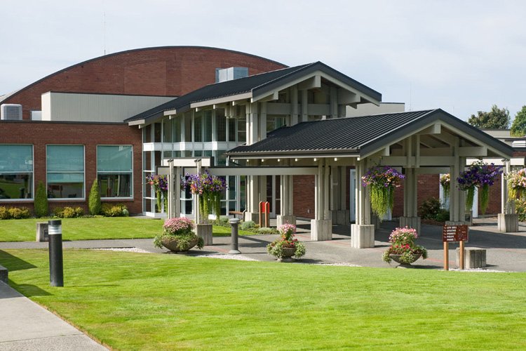 A modern building with a curved red brick roof, glass entrance, and a covered drop-off area with hanging flower baskets, surrounded by a landscaped lawn.