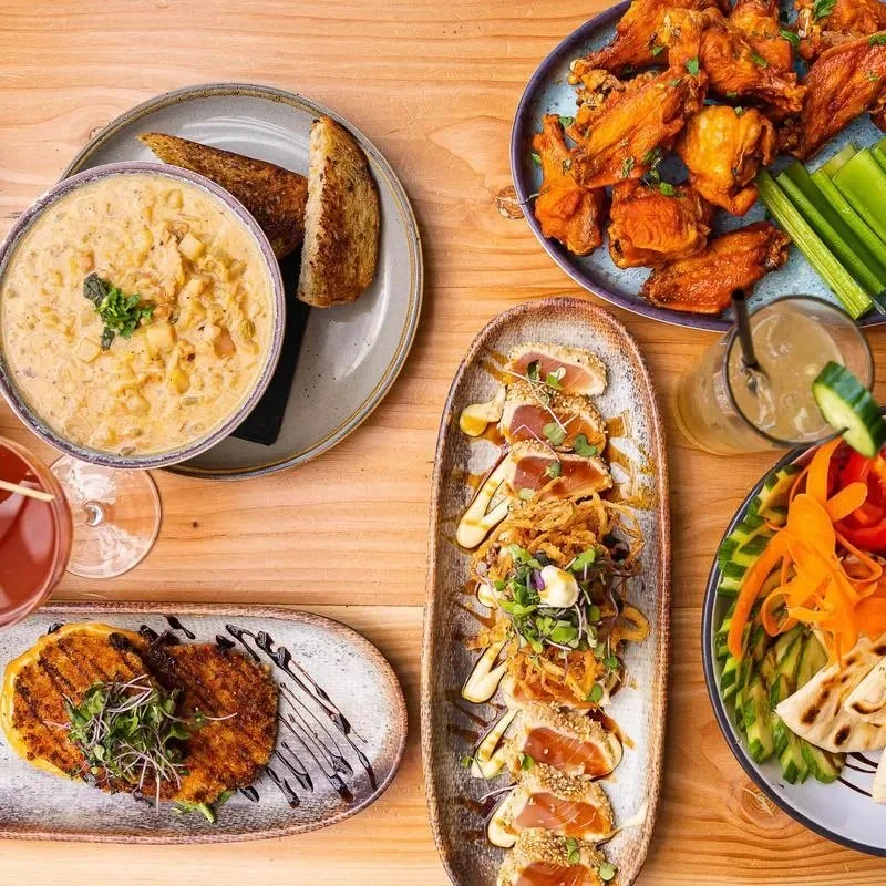 Assorted dishes including a bowl of potato soup with toast, buffalo wings with celery sticks, a plate of sushi rolls with sauce, a grilled breaded cutlet with sauce and microgreens, fresh veggie salad with carrots and cucumber, and cocktails on a wooden table.