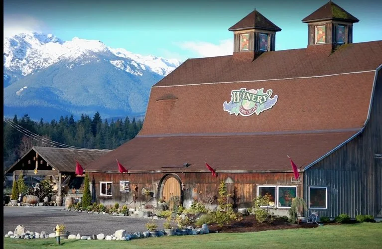 A rustic winery building with a large sloped roof, two small towers, and a mountain range with snow-capped peaks in the background.