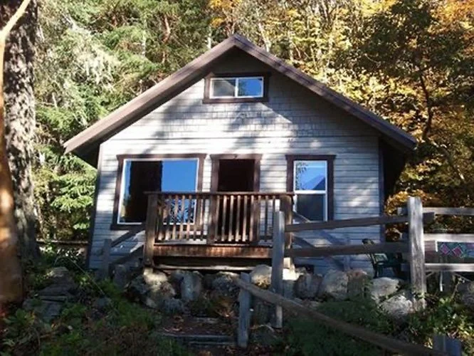 A small wooden house with a pitched roof, located in a forested area, featuring a front door, windows, and a small balcony.