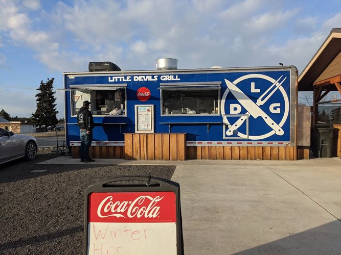 A blue food truck named Little Devils Grill with a DLG logo on the side. A person stands near the window, and a Coca-Cola sign is on a stand in the foreground. The parking lot and a building are visible in the background.
