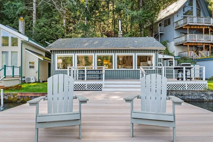 A wooden dock with two Adirondack chairs facing a lakeside house with large windows and a deck, surrounded by trees.