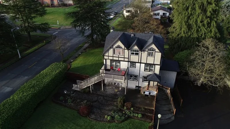 A large, multi-story house with Tudor-style architecture, surrounded by a garden and trees, with a deck and stairs leading to a backyard.