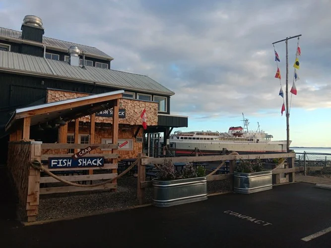 Exterior view of a seafood restaurant called Fish Shack with a pier and boat in the background, and nautical flags flying on a pole.