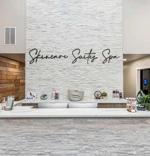 Front desk of a spa with a white brick wall, a sign reading 'Skincare Suites Spa,' and decorative items including a face sculpture, plants, and skincare products.