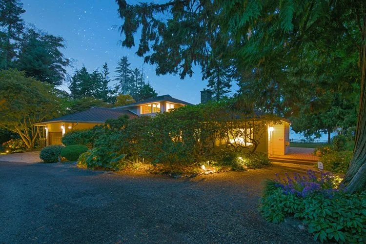 A house illuminated with warm outdoor lighting, surrounded by lush trees and bushes, during dusk or evening.