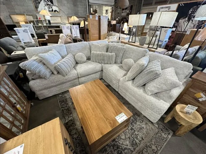 A large, light gray sectional sofa with various throw pillows and round bolster pillows in a furniture store. A wooden coffee table is in front of the sofa, with other furniture and lighting fixtures visible in the background.
