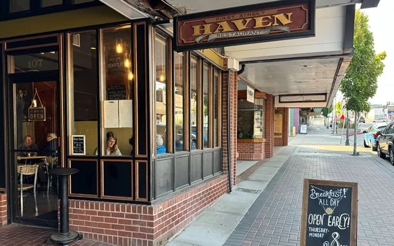 Exterior of First Street Haven Restaurant with a black and gold sign, large windows revealing people dining inside, and a sidewalk chalkboard advertising breakfast served all day.